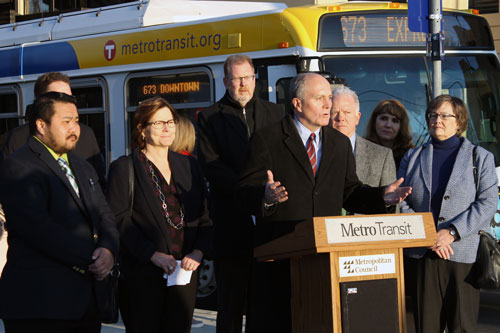 A man speaking at a podium with people and a bus in the background.