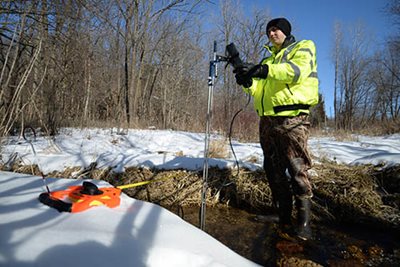 A person in a bright green jacket standing in a creek with measuring equipment in winter.