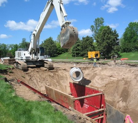 Image of previous sewer construction project in process, showing an excavator on the edge of a hole with a construction structure at the bottom.  The excavator is lowering a piece of pipe into the hole.