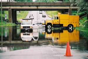 Two trucks in standing water in a paved road under an overpass.